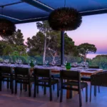 Longue table dressée sous la pergola de l'Hôtel Villa Padulaccia au crépuscule, suspensions noires et pins