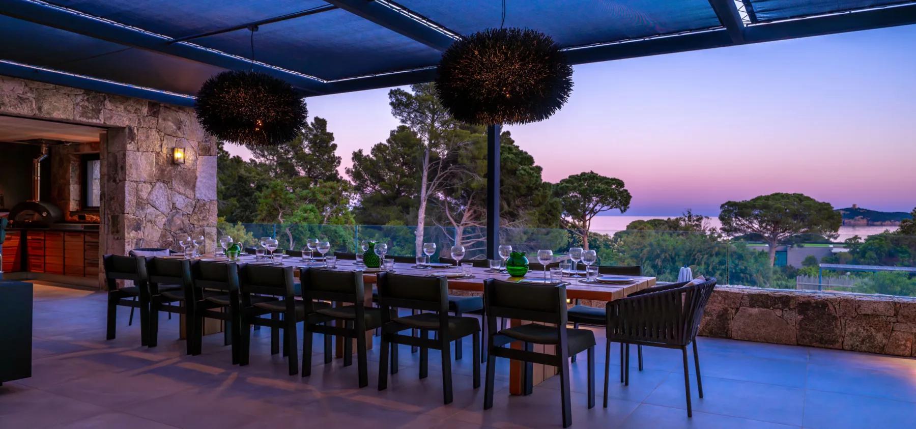 Longue table dressée sous la pergola de l'Hôtel Villa Padulaccia au crépuscule, suspensions noires et pins
