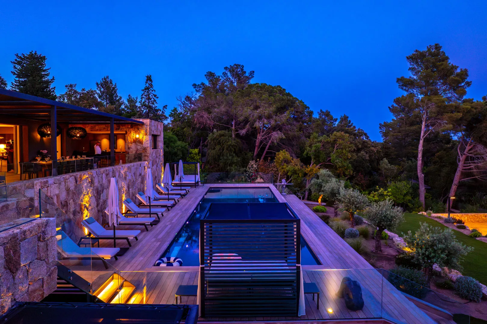Piscine de l'Hôtel Villa Padulaccia au crépuscule, transats le long de la terrasse éclairée et pins alentour
