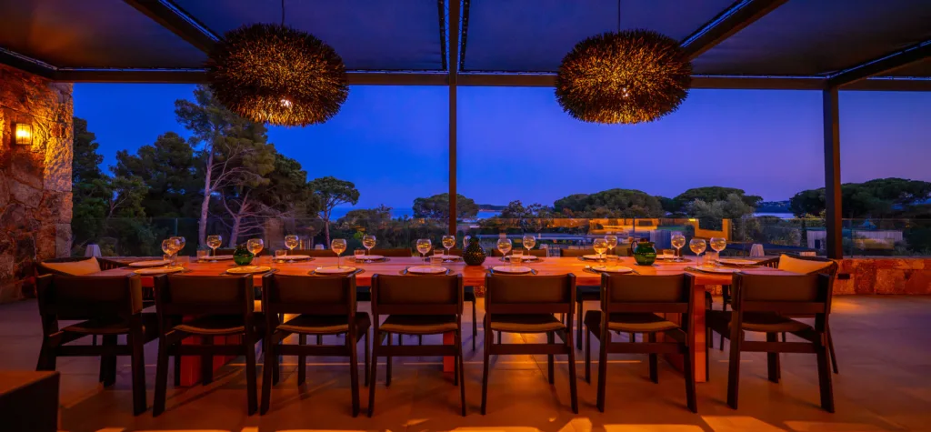 Longue table dressée sous la pergola de l'Hôtel Villa Padulaccia à l'heure bleue, verres alignés et suspensions allumées