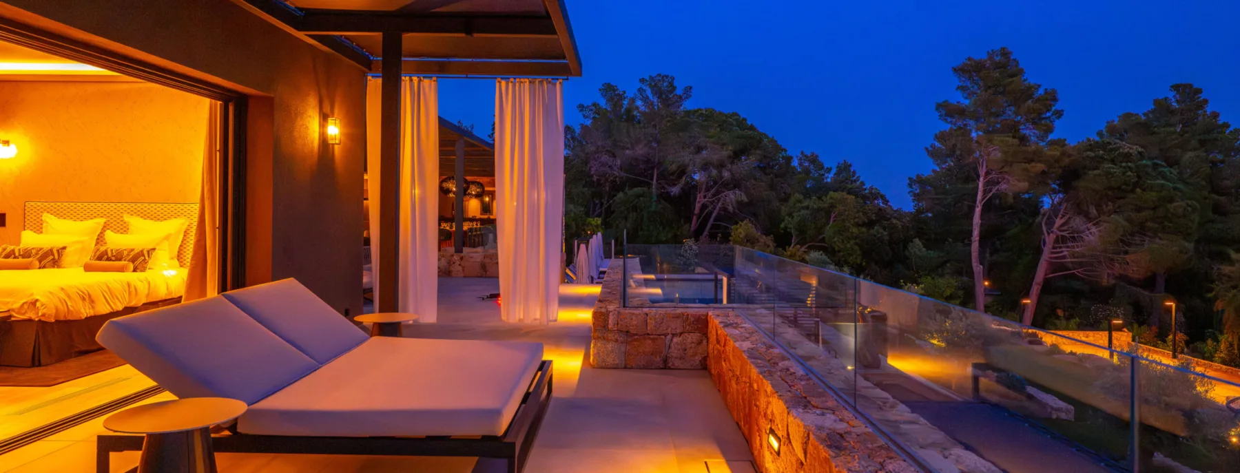 Chambre éclairée et lit de repos sur la terrasse de l'Hôtel Villa Padulaccia à la nuit tombée, rideaux blancs