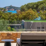 Lit de repos et table d'appoint sur une terrasse de l'Hôtel Villa Padulaccia, tour génoise et mer au-delà des pins