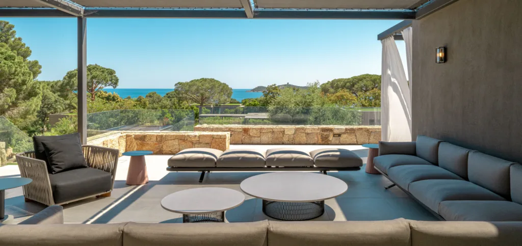 Salon extérieur couvert de l'Hôtel Villa Padulaccia, canapés gris et tables basses face aux pins et à la mer