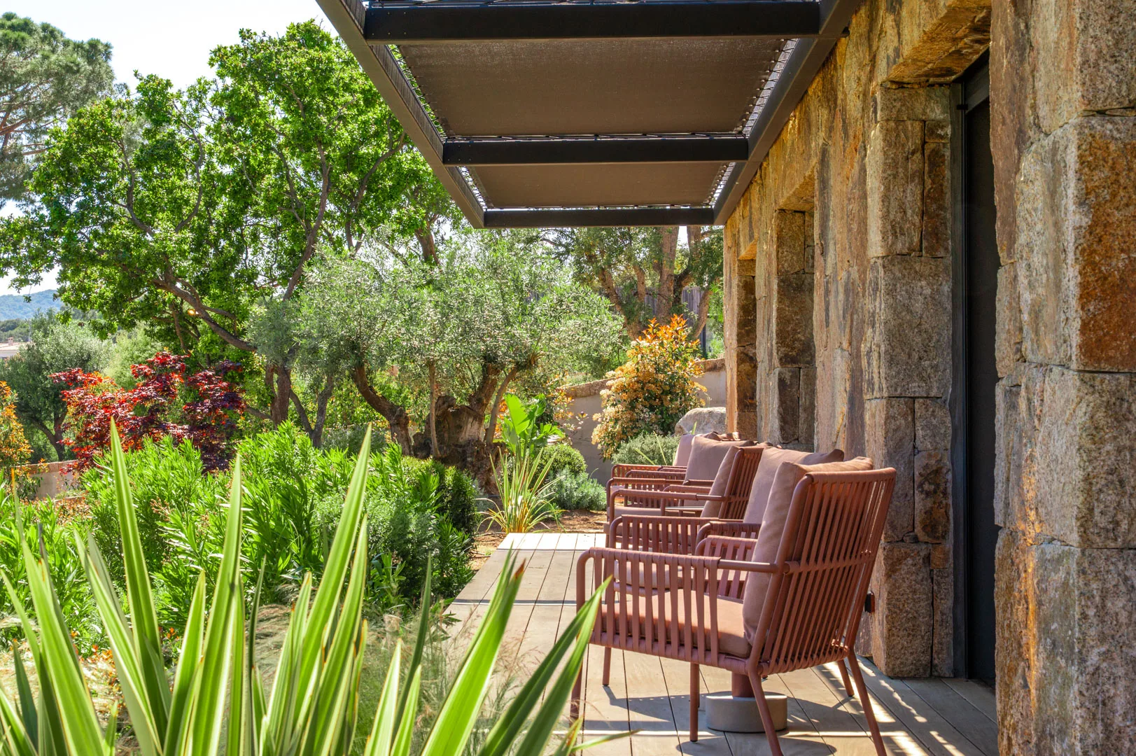 Terrasse en bois au pied d'un mur de pierre, fauteuils roses face au jardin de l'Hôtel Villa Padulaccia